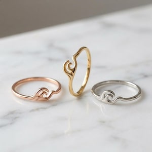 Op de afbeelding: Drie dunne metalen ringen, elk voorzien van een klein, gestileerd golfontwerp. Eén ring is roségoudkleurig, één is goudkleurig en één is zilverkleurig. De ringbanden zijn glad en rond, waarbij het golfmotief in de band is verwerkt.