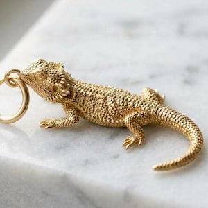 Könnte beinhalten: Ein goldfarbener Metallanhänger in Form einer Bartagame mit detaillierten Schuppen und einem strukturierten Körper. Die Eidechse ist in einer Ruheposition dargestellt und verfügt über einen runden Biegering am Kopf zum Aufhängen.