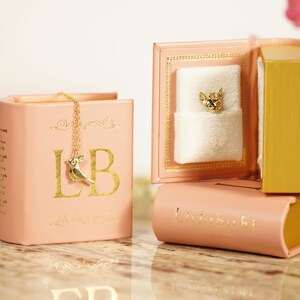 A pink book-shaped jewelry box with a gold chain and a small white charm in the front, placed on a marble surface.