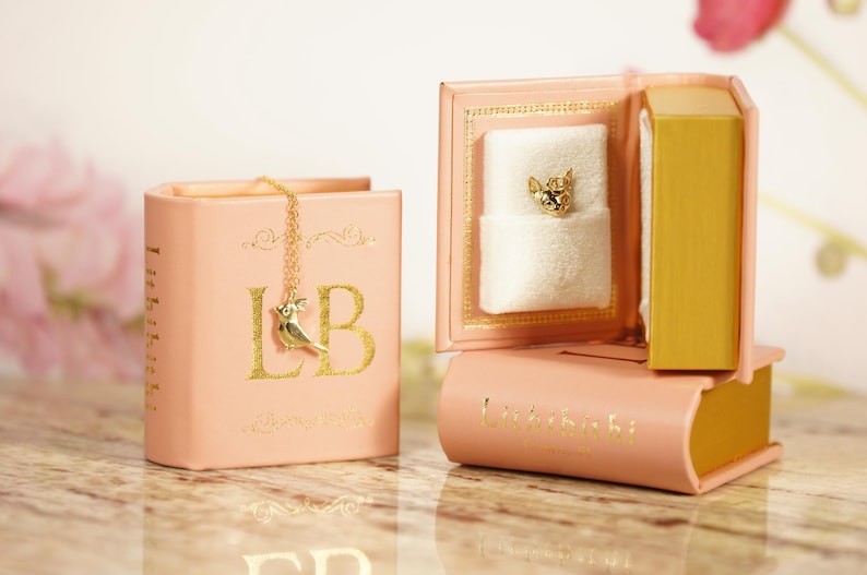 A pink book-shaped jewelry box with a gold chain and a small white charm in the front, placed on a marble surface.