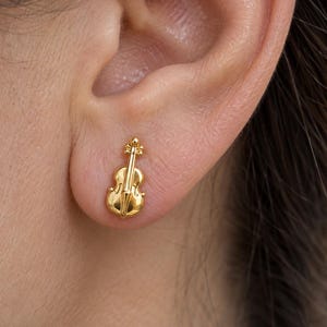 Könnte beinhalten: Ein kleiner goldfarbener Ohrstecker in Form einer Geige. Der Ohrstecker hat eine polierte metallische Oberfläche mit detaillierten Gravuren, die den Korpus, den Hals und die Schnecke des Saiteninstruments darstellen und direkt am Ohrläppchen anliegen.