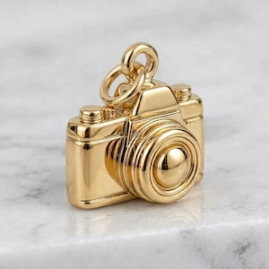 Könnte beinhalten: Ein glänzender goldfarbener Metallanhänger in Form einer Vintage-Kamera. Der Anhänger verfügt über ein markantes kreisförmiges Objektiv, einen rechteckigen Körper und einen kleinen Auslöser an der Oberseite. Ein Ring ist oben befestigt.
