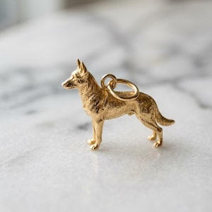 May include: A gold-toned metal charm shaped like a German Shepherd dog standing in profile. The charm features a textured surface to mimic fur and includes a small circular jump ring attached to the back for hanging on a chain or bracelet.