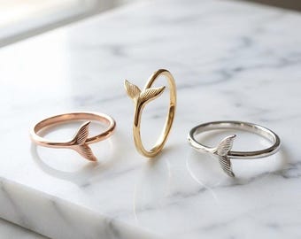 May include: Three rings featuring a whale tail or mermaid fin design. One ring is rose gold, one is yellow gold, and one is silver. Each ring has a thin, smooth band with the fin motif extending upward from the point where the band ends meet.