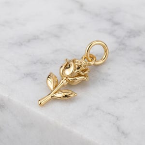 May include: A polished gold-tone metal pendant shaped like a rose flower with a single stem and two leaves. The pendant features a circular jump ring at the top for attachment, showcasing a smooth, reflective surface with detailed petal contours.