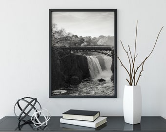 May include: Black and white photograph of a waterfall with a bridge over it. The photo is framed in a black frame and is hanging on a white wall.