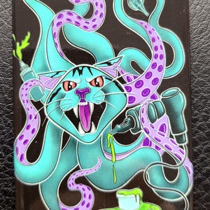 May include: A rectangular magnet featuring a cartoon cat with teal fur and red eyes, combined with purple tentacles. The cat has an open mouth with a pink tongue and sharp teeth. The tentacles are wrapped around tattoo guns. The words "Shatterproof Tattoo" are at the bottom.