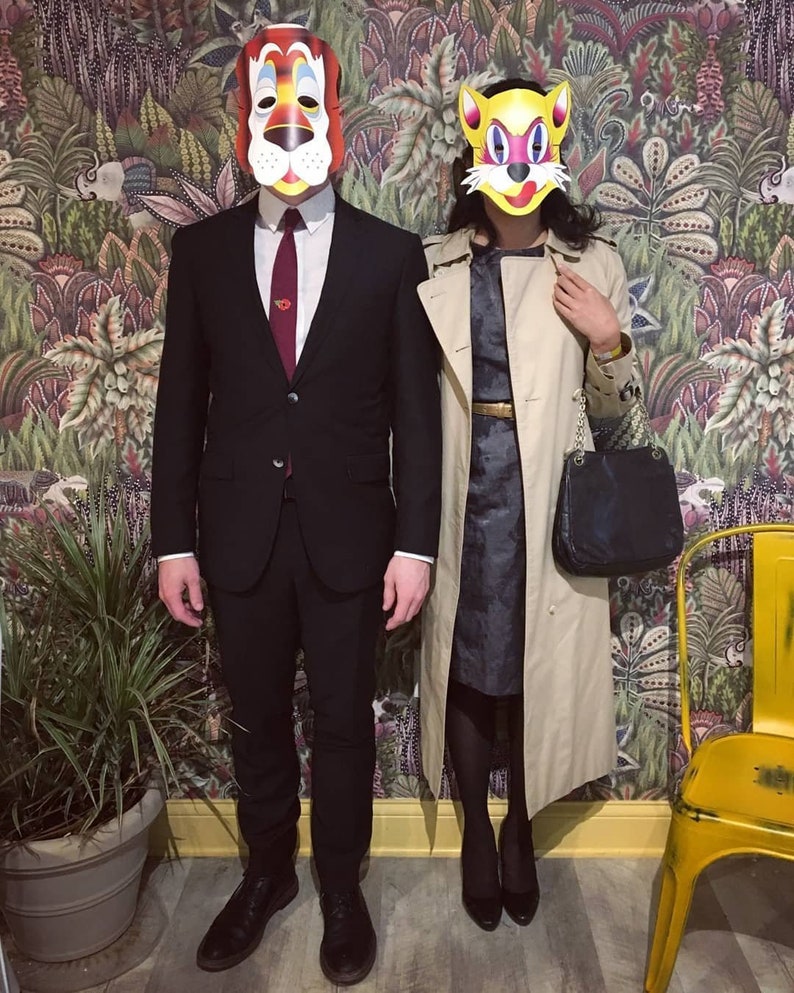 Puede incluir: Una pareja con m&aacute;scaras de animales, un tigre y un gato, est&aacute;n de pie frente a una pared con patrones. El hombre lleva un traje negro y una corbata roja. La mujer lleva una gabardina beige sobre un vestido azul oscuro.