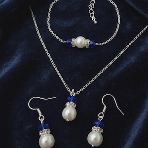 May include: A jewellery set consisting of a necklace, a bracelet, and a pair of drop earrings. Each piece features a white faux pearl, a faceted royal blue bead, and a silver-toned spacer ring embedded with small clear crystals on a silver chain.