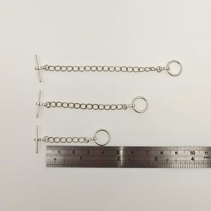 May include: Three silver-colored chain extenders with a T-bar clasp on one end and a ring on the other. A ruler is placed below the extenders to show the length of the chains. The chains are approximately 5 inches, 4 inches, and 3 inches long.