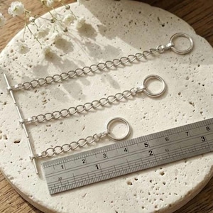 May include: Three silver-toned metal chain extenders with circular rings at one end and T-bar toggles at the other. A metal ruler is placed alongside the chains, showing the longest chain measures approximately 3.5 inches in total length.