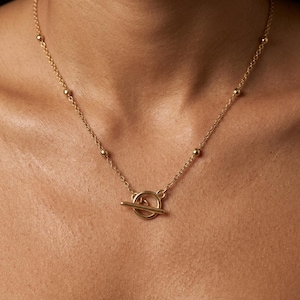 Peut inclure: Un collier à chaîne doré orné d'une série de petites perles sphériques régulièrement espacées le long des maillons. Le collier est fixé par un fermoir à bascule circulaire et une barre horizontale positionnée au centre du cou.