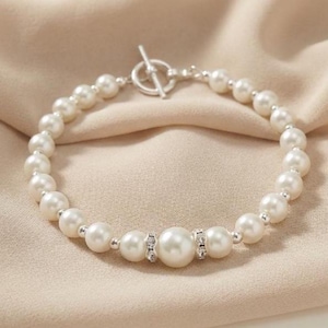 May include: A pearl bracelet with a silver clasp. The bracelet features round, white pearls strung together with silver accents. A larger pearl is centered, flanked by smaller decorative stones.