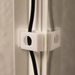 Może przedstawiać: Białe plastikowe zaciski kablowe przymocowane do szklanej powierzchni, utrzymujące czarny kabel na miejscu.