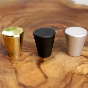 May include: Three cabinet knobs in gold, black, and silver. The knobs are round and have a cone shape. They are displayed on a wooden surface.