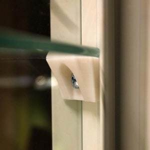 Può includere: Una staffa per mensole in plastica bianca con una vite fissata. La staffa è montata su una cornice bianca.