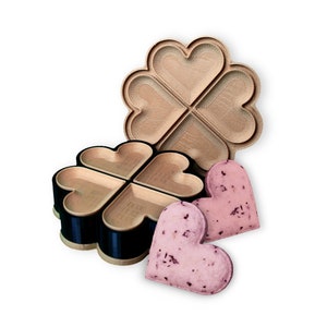 Könnte beinhalten: Ein hölzerner herzförmiger Waffeleisen mit vier Abschnitten, neben zwei herzförmigen Waffeln. Das Waffeleisen ist zweiteilig, mit einem schwarzen und goldenen Sockel. Die Waffeln sind rosa mit dunklen Flecken.