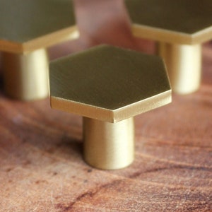 Könnte beinhalten: Drei goldene sechseckige Schrankknöpfe mit glatter, polierter Oberfläche. Die Knöpfe sind in einer Reihe auf einer Holzoberfläche angeordnet.