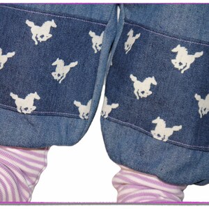 Pu&ograve; includere: Un paio di pantaloni di jeans blu con disegni di cavalli bianchi sui polsini. I pantaloni sono arrotolati in fondo, rivelando un tessuto a righe rosa e bianco sotto.