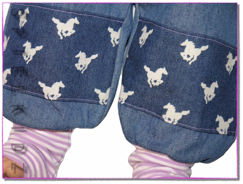 Pu&ograve; includere: Un paio di pantaloni di jeans blu con disegni di cavalli bianchi sui polsini. I pantaloni sono arrotolati in fondo, rivelando un tessuto a righe rosa e bianco sotto.