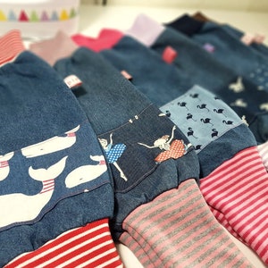 Pu&ograve; includere: Una collezione di pantaloni di jeans con diverse toppe di tessuto stampato colorato e polsini a righe. I pantaloni presentano disegni di balene, ballerine e fenicotteri.