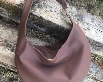 May include: A brown, crescent-shaped shoulder bag with a long strap. The bag is made of a smooth material and features a gold-colored clasp. The bag is lying on a weathered wooden surface.