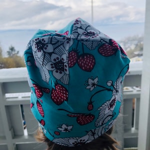 Może przedstawiać: Turkusowa czapka z nadrukiem białych i czerwonych truskawek. Czapka wykonana jest z miękkiej, elastycznej tkaniny.