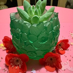May include: A green glass planter with a floral design, holding a green succulent plant. Red fabric flowers are scattered around the base of the planter on a pink surface. The planter is approximately 10 cm in diameter.