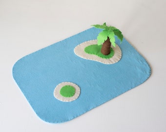 Puede incluir: Una escena de isla de fieltro con un océano azul claro y dos islas verdes. Una isla presenta un árbol marrón con hojas verdes. La escena está sobre un fondo blanco.