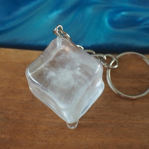 Può includere: Un portachiavi a forma di cubetto di ghiaccio trasparente con un anello portachiavi in metallo argentato. Il cubetto di ghiaccio ha una forma quadrata con bordi arrotondati e presenta una piccola goccia nella parte inferiore.