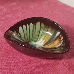 Könnte beinhalten: Eine dunkelbraune, dreieckige Keramikschale mit glänzendem Finish. Das Innere zeigt ein gemaltes Design mit grünen, gelben und blauen Farbtönen. Die Schale steht auf einer rosa Tischdecke.