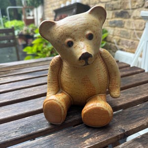 May include: A ceramic teddy bear figurine with a tan and brown speckled finish. The bear has dark brown eyes and a small nose. The bear is sitting on a wooden table.