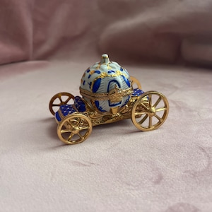 Op de afbeelding: Een decoratieve sieradendoos in de vorm van een koets. De doos heeft een goudkleurig metalen frame met blauwe en witte emaille details. De wielen zijn goudkleurig en de bovenkant van de koets is versierd met blauwe en gouden bloemmotieven.
