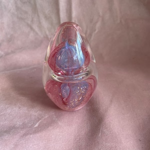 May include: A clear glass paperweight with a pink and blue floral design. The interior features a swirling pattern of pink and blue, with gold flecks. The paperweight has an oval shape.