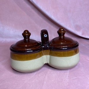 May include: A ceramic condiment server with two connected compartments, each with a lid. The server is brown, tan, and cream-coloured, with a handle for easy carrying. The lids have a decorative knob.