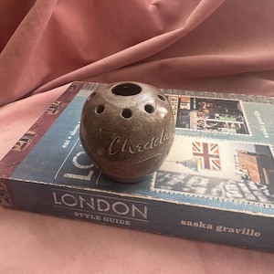 May include: A speckled brown ceramic candle holder with multiple holes. The word "Cheddar" is written on the side in a cursive font. The candle holder sits on a book with the words "London Style Guide".