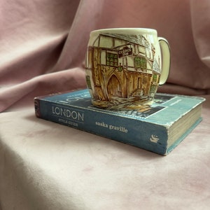 May include: A vintage ceramic mug featuring a detailed illustration of the George Inn, with a handle and cream-colored rim. The mug rests on a blue book titled "LONDON STYLE GUIDE" by saska graville.