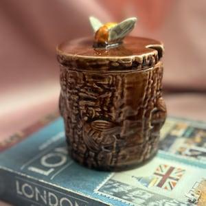 Peut inclure: Un pot en céramique marron avec un couvercle et une poignée en forme d'abeille. Le pot a un motif texturé de tronc d'arbre. Le pot est posé sur un livre avec le mot "LONDON" sur la couverture.