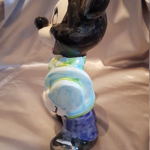 Op de afbeelding: Een keramieken beeldje van Mickey Mouse, met zwarte oren en hoofd, en een wit gezicht. Mickey draagt een lichtblauwe jas met een groen en wit patroon, een paarse broek en zwarte schoenen. Het beeldje staat op een satijnen achtergrond.
