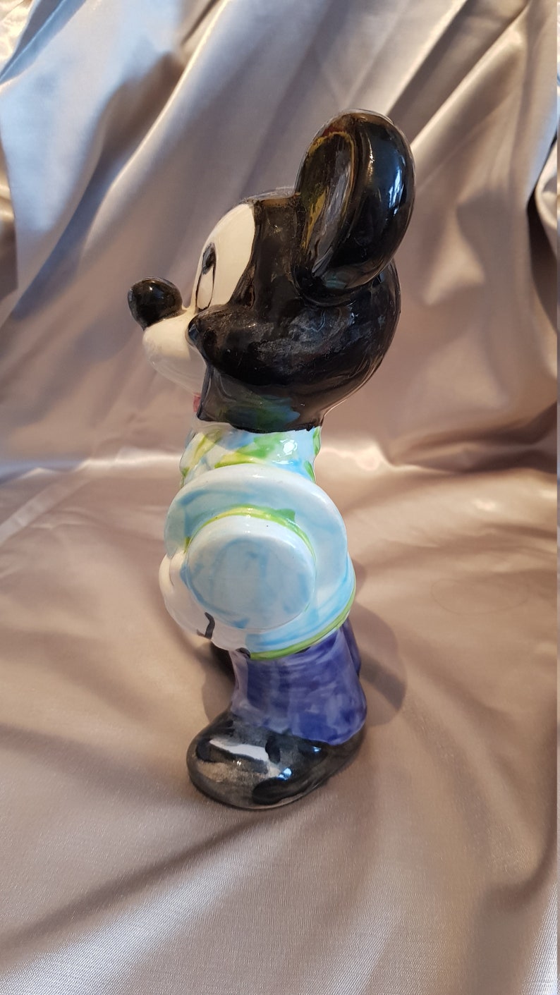 Op de afbeelding: Een keramieken beeldje van Mickey Mouse, met zwarte oren en hoofd, en een wit gezicht. Mickey draagt een lichtblauwe jas met een groen en wit patroon, een paarse broek en zwarte schoenen. Het beeldje staat op een satijnen achtergrond.