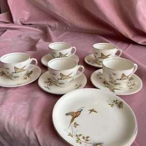 Op de afbeelding: Een vintage theeservies met zes kopjes met schotels en drie borden. De witte porseleinen kopjes en borden zijn versierd met een fazantenmotief in bruin, blauw en goud. De randen van de kopjes en borden zijn goudkleurig.
