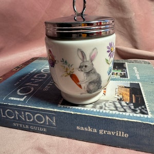 May include: A white ceramic egg cup with a silver lid and handle. The cup is decorated with a gray rabbit holding a carrot, a butterfly, and flowers. The egg cup is sitting on a blue book.