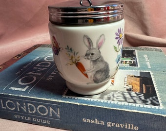 May include: A white ceramic egg cup with a silver lid and handle. The cup is decorated with a gray rabbit holding a carrot, a butterfly, and flowers. The egg cup is sitting on a blue book.
