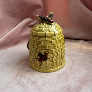 May include: A ceramic honey pot shaped like a beehive, with a lid and a bee-shaped handle. The pot is yellow-green with a textured design and painted bees on the side. The background is a pink fabric.