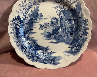 Op de afbeelding: Witte keramische bord met een geschulpte rand, met een blauw-wit pastorale scène. Het ontwerp omvat een paardenkoets, gebouwen en bomen. De tekst onderaan luidt "Down to the Smithy".