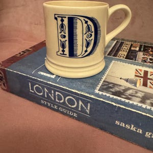 May include: Cream-colored ceramic mug with a large, ornate blue letter "D". The mug has a handle and sits on a blue book titled "London Style Guide". The mug's design features floral accents.