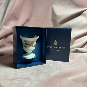 May include: A white porcelain egg cup with a colorful bird and floral design, presented in a navy blue box. The box is labeled "ROYAL WORCESTER BONE CHINA". The egg cup has a pedestal base and a flared top.
