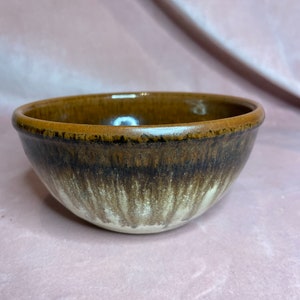 以下が含まれることがあります： 茶色とクリーム色の釉薬を施した、手作りの小さなセラミックボウル。 ボウルは丸みを帯びた形状で、縁がわずかに広がっています。 釉薬は縁の濃い茶色から底のクリーム色に変化し、独特の視覚効果を生み出しています。