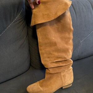 May include: A tall, brown suede boot with a folded-over top. The boot has a low heel and a slouchy design. The boot is made of a soft, textured material and appears to be unworn. The boot is resting on a dark gray surface.
