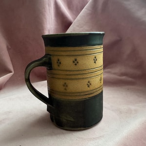 Könnte beinhalten: Eine Keramik-Tasse mit schwarzem Rand und Boden sowie einem beigefarbenen Mittelteil. Der beigefarbene Abschnitt weist ein sich wiederholendes Muster aus kleinen, vierblättrigen Blumendesigns auf. Der Griff ist dunkelgrau.